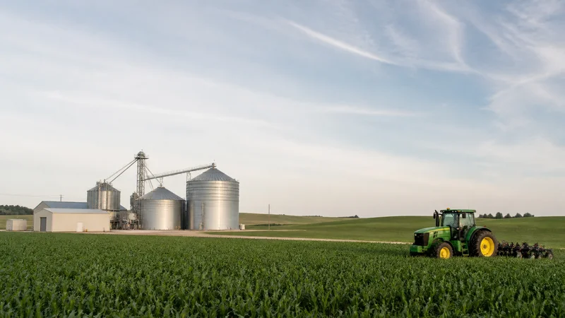 Wide establishing view of a large farm fields with a tractor and grain silos, soft daylight, open sky above for overlaid text