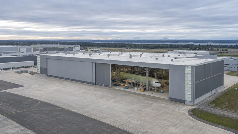 High aerial of a aircraft final-assembly hangar with a fuselage section visible through the open doors, overcast soft light, uncluttered ground and sky bands