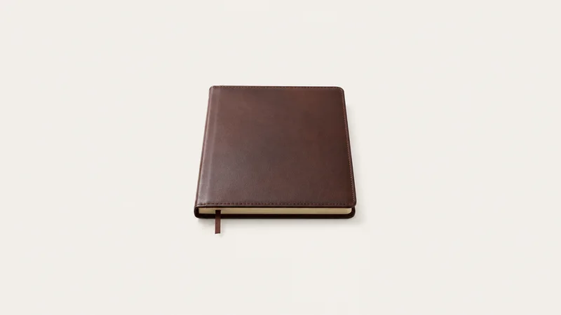 Isolated a leather-bound blank portfolio ledger with a ribbon marker centred on a seamless pale off-white studio background, generous empty margin on every side for overlaid branding