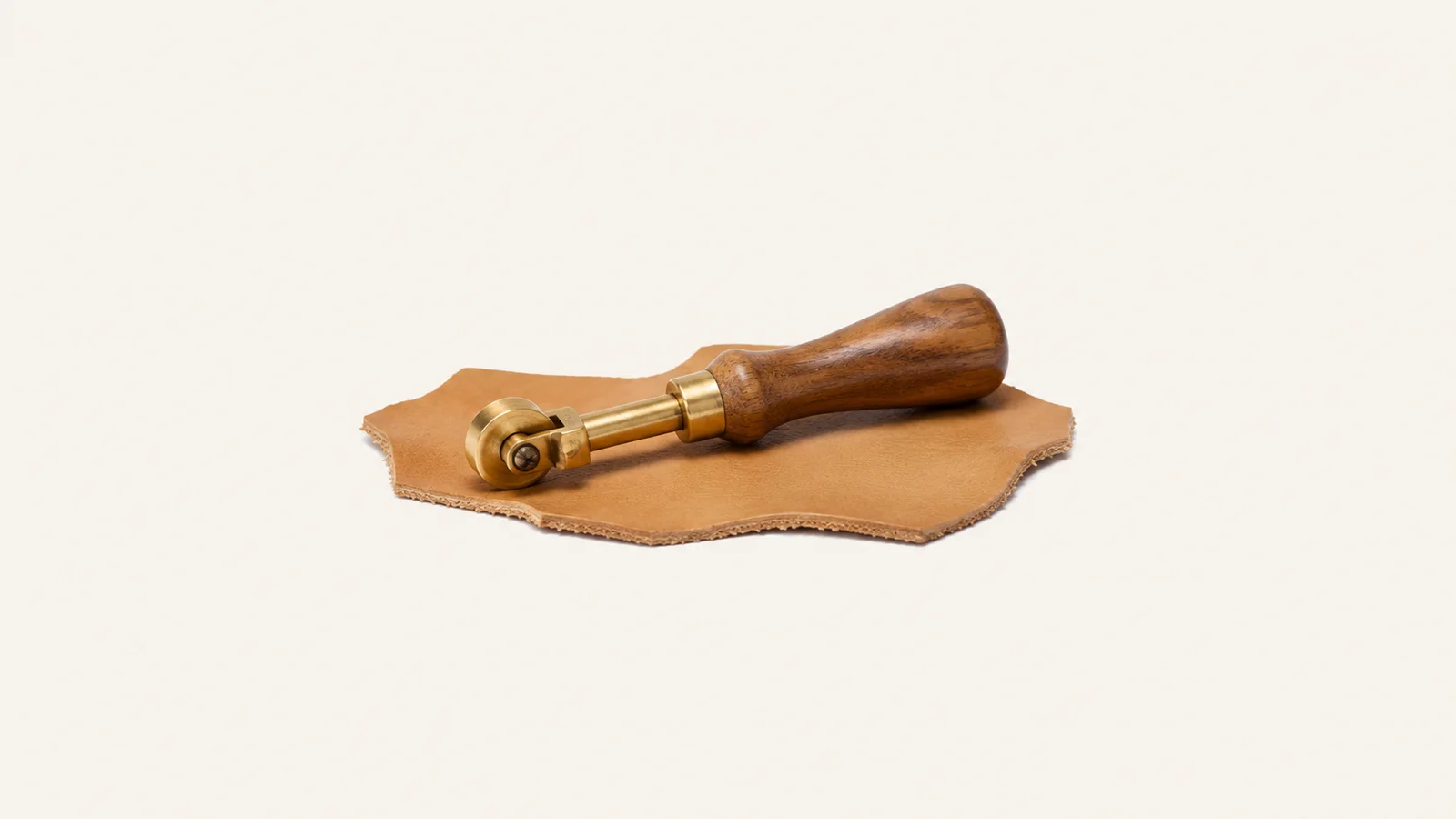 Isolated a brass edge creaser with a wooden handle on a scrap of tan hide centred on a seamless pale off-white studio background, generous empty margin on every side for overlaid branding