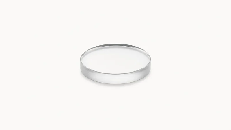 Isolated an uncoated glass lens blank with a ground edge centred on a seamless pale off-white studio background, generous empty margin on every side for overlaid branding