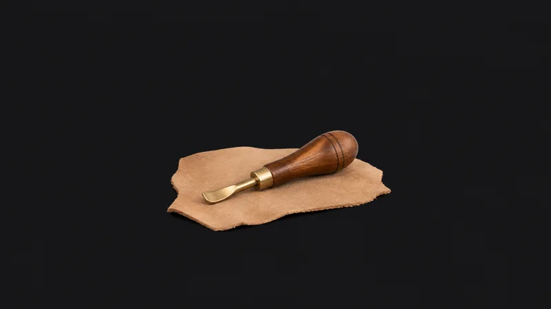 Isolated a brass edge creaser with a wooden handle on a scrap of tan hide centred on a seamless deep charcoal near-black studio background, generous empty margin on every side for overlaid branding
