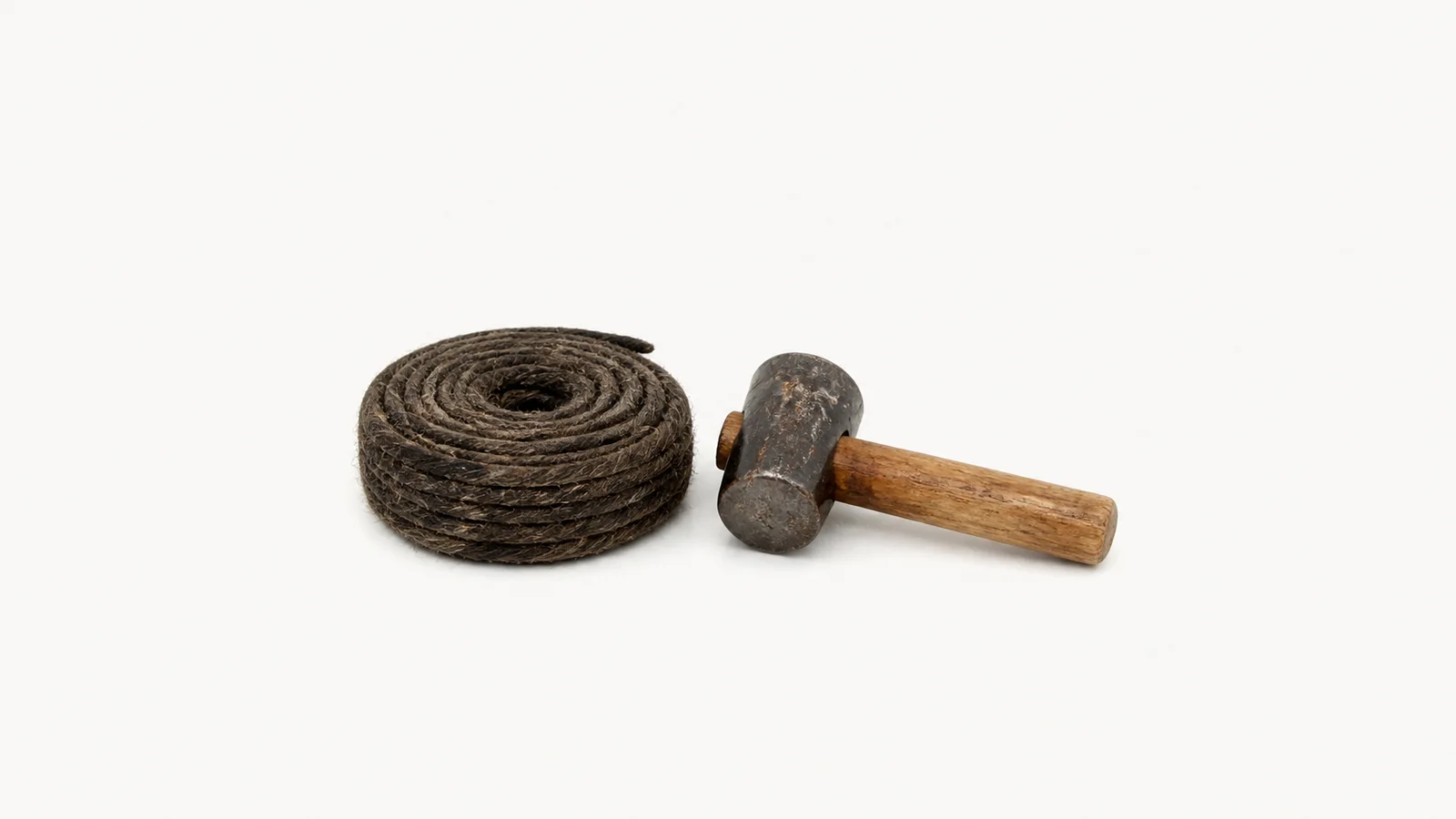 Isolated a coil of tarred hemp caulking cotton beside a caulking mallet centred on a seamless pale off-white studio background, generous empty margin on every side for overlaid branding