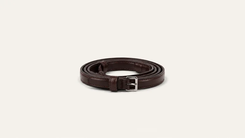 Isolated a coiled leather rein with a buckle billet centred on a seamless pale off-white studio background, generous empty margin on every side for overlaid branding