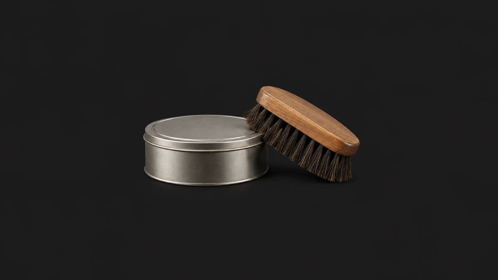 Isolated a tin of paste wax with a coloured horsehair polishing brush centred on a seamless deep charcoal near-black studio background, generous empty margin on every side for overlaid branding