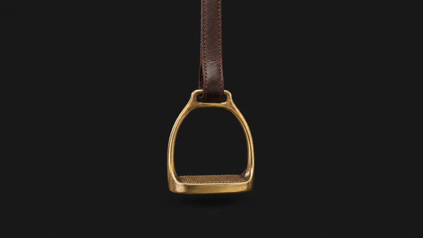 Isolated a brass stirrup iron on a leather stirrup strap centred on a seamless deep charcoal near-black studio background, generous empty margin on every side for overlaid branding