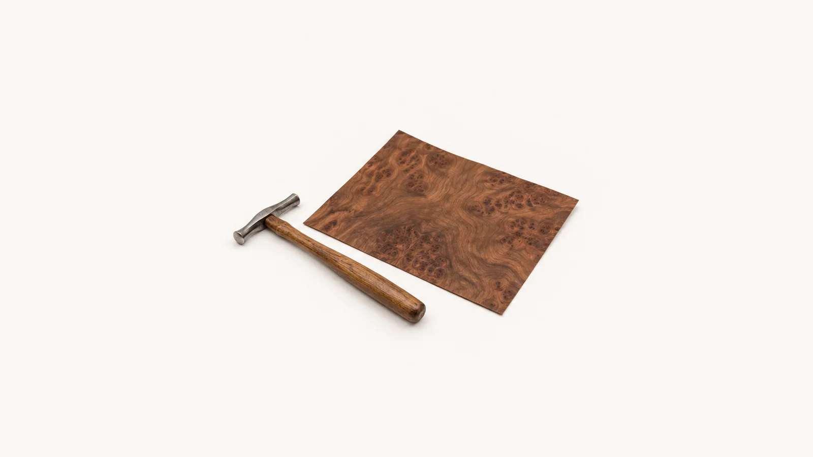 Isolated a veneer hammer beside a sheet of burr-walnut veneer centred on a seamless pale off-white studio background, generous empty margin on every side for overlaid branding