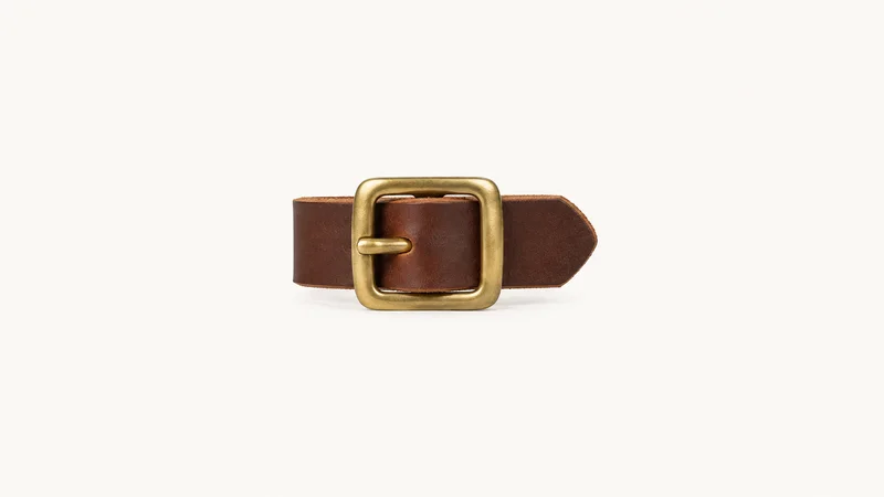 Isolated a brass belt buckle on a strip of unstitched bridle leather centred on a seamless pale off-white studio background, generous empty margin on every side for overlaid branding