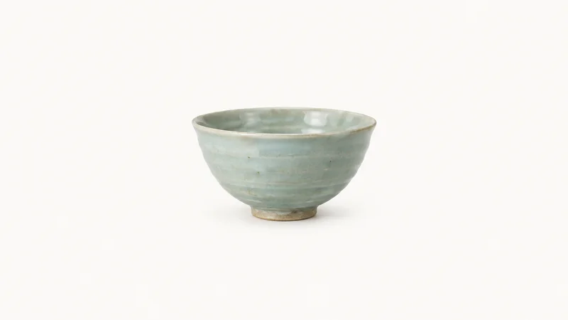 Isolated a hand-thrown porcelain bowl with a celadon glaze centred on a seamless pale off-white studio background, generous empty margin on every side for overlaid branding