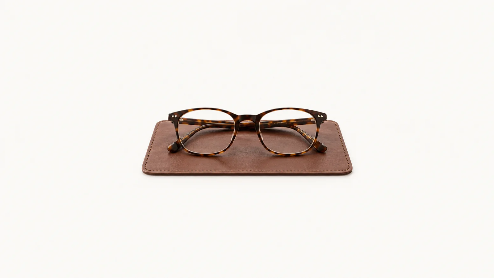 Isolated a tortoiseshell-framed pair of reading spectacles on a leather blotter centred on a seamless pale off-white studio background, generous empty margin on every side for overlaid branding