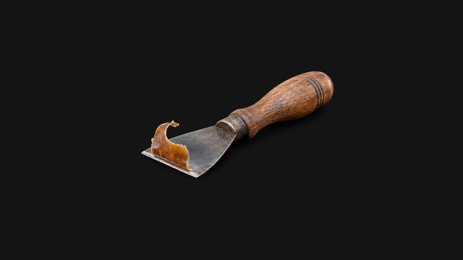 Isolated a wooden hand scraper with a curled shaving of old varnish centred on a seamless deep charcoal near-black studio background, generous empty margin on every side for overlaid branding
