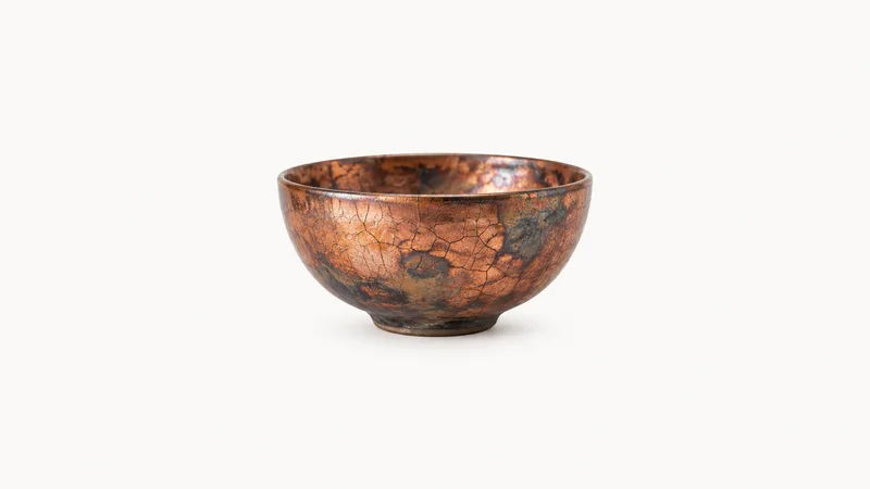 Isolated a raku-fired bowl with a crackled copper glaze centred on a seamless pale off-white studio background, generous empty margin on every side for overlaid branding