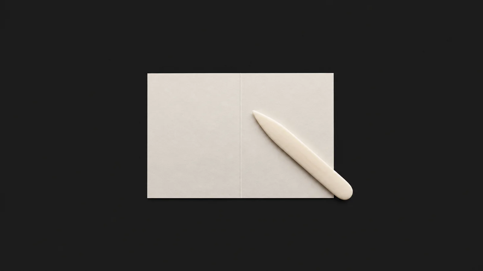 Isolated a bone folder and a scored sheet of card centred on a seamless deep charcoal near-black studio background, generous empty margin on every side for overlaid branding