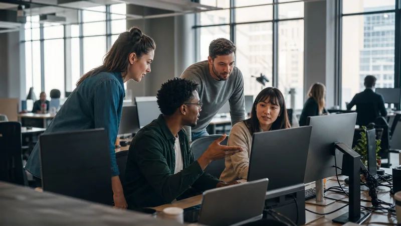 A few software engineers working together, open-plan engineering floor in the background, bright daylight through the windows