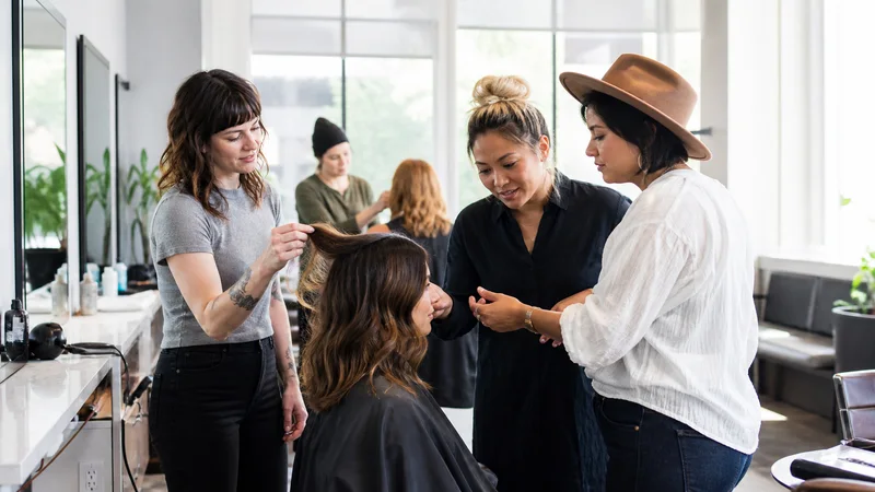 A few stylists working together, bright salon styling stations in the background, bright daylight through the windows