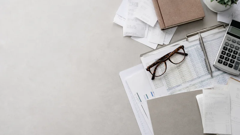 Styled flat-lay of spreadsheets, receipts and glasses on a clean background, clean even studio light, generous negative space for overlaid text