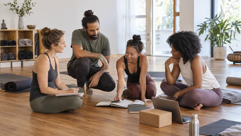 A few yoga instructors working together, open wood-floor studio with rolled mats in the background, soft even indoor light